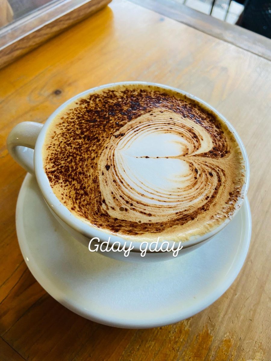 A cappuccino in a white cup and saucer on a timber table, with a heart poured into the crema and a dusting of chocolate.