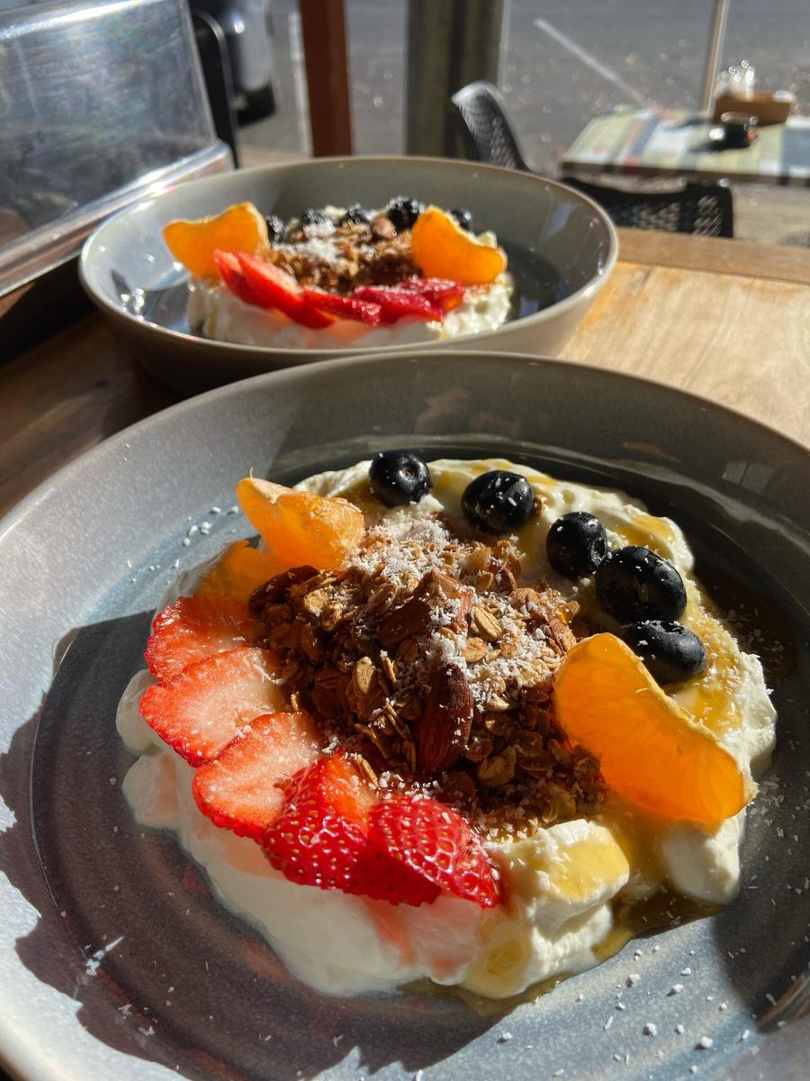 A granola bowl in morning window light: thick yoghurt topped with toasted granola, almonds, desiccated coconut, sliced strawberry, blueberries and mandarin segments, with honey pooling at the edge.