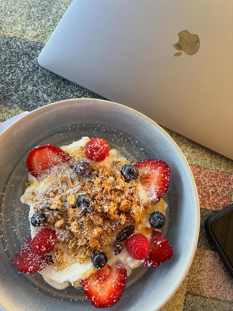 A granola bowl with strawberries, raspberries, blueberries and coconut on a speckled ceramic plate, sitting beside a closed laptop and a phone on a tiled table.