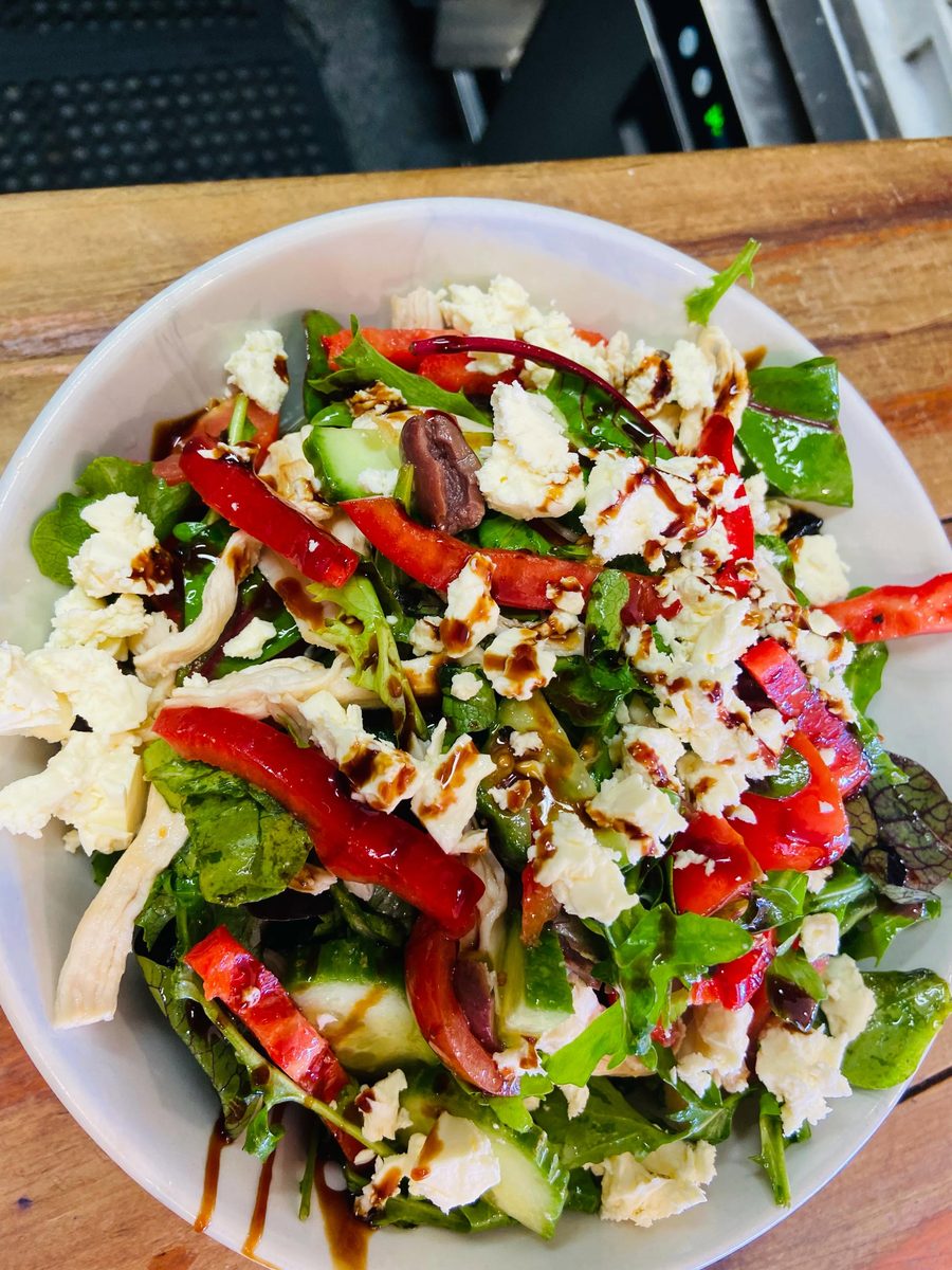 A Greek salad in a white bowl: mixed leaves, cucumber, roasted red capsicum, kalamata olives and crumbled feta with shredded chicken, finished with a balsamic drizzle.